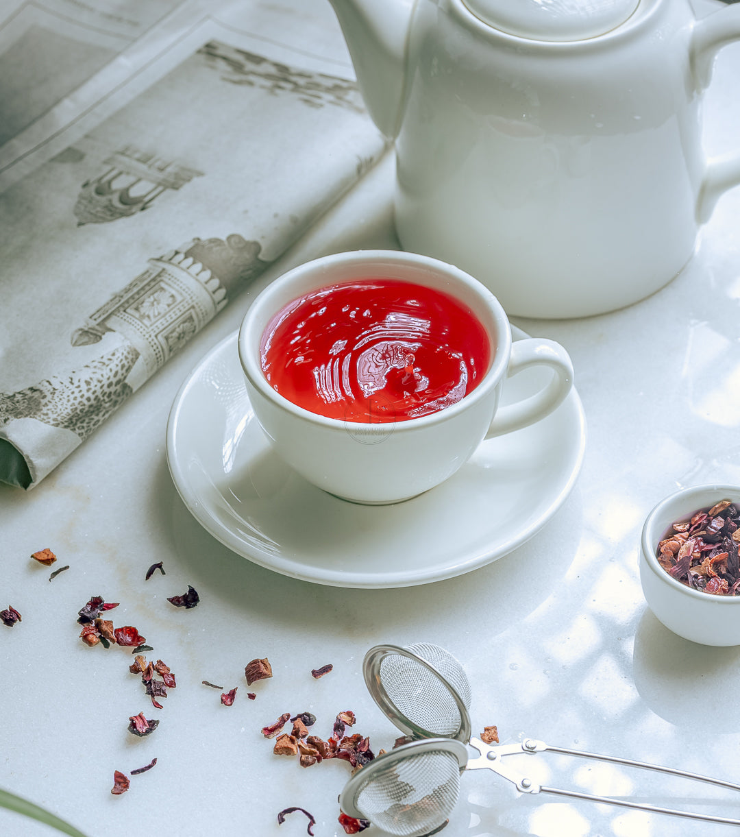 A cup of Loafology Bakery & Café's Strawberry & Kiwi Tea sits gracefully on a saucer beside a teapot, surrounded by loose tea leaves and a tea strainer on a white table near a newspaper. This delightful nighttime beverage provides relaxation and an invigorating boost of Vitamin C to help you unwind.