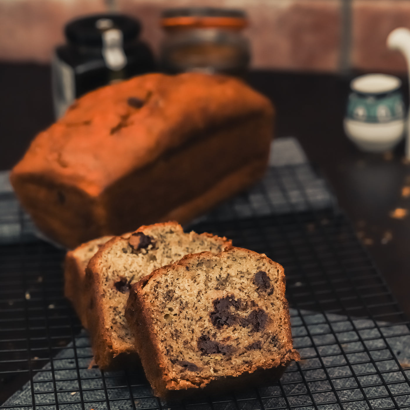 A loaf of Banana Bread from Loafology Bakery & Café, glistening with a hint of butter, is placed on a cooling rack, with three slices cut from it. Containers and a cup are in the blurry background.