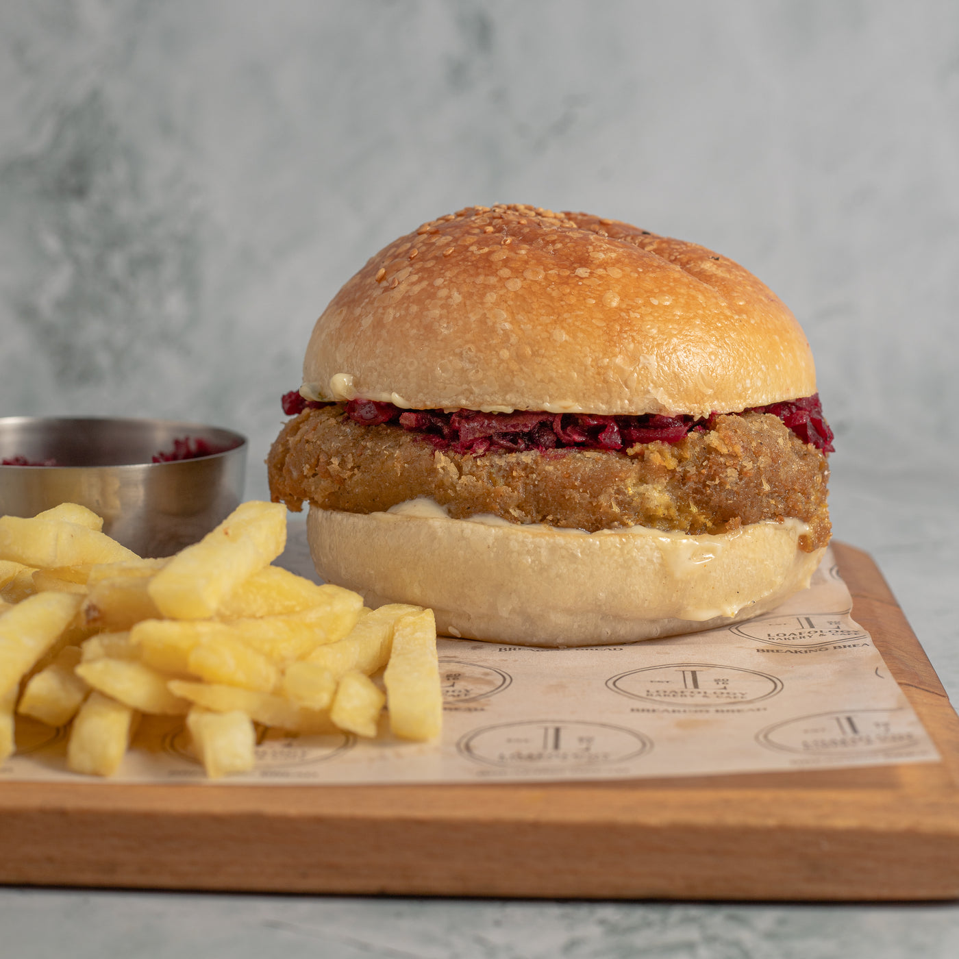 Introducing the Golden Vegetarian Burger from Loafology Bakery & Café: a flavorful breaded tofu patty nestled in a sesame seed bun, topped with beetroot slaw and creamy mayonnaise, all served on a rustic wooden tray alongside crispy fries and a small dish of our signature homemade salsa.