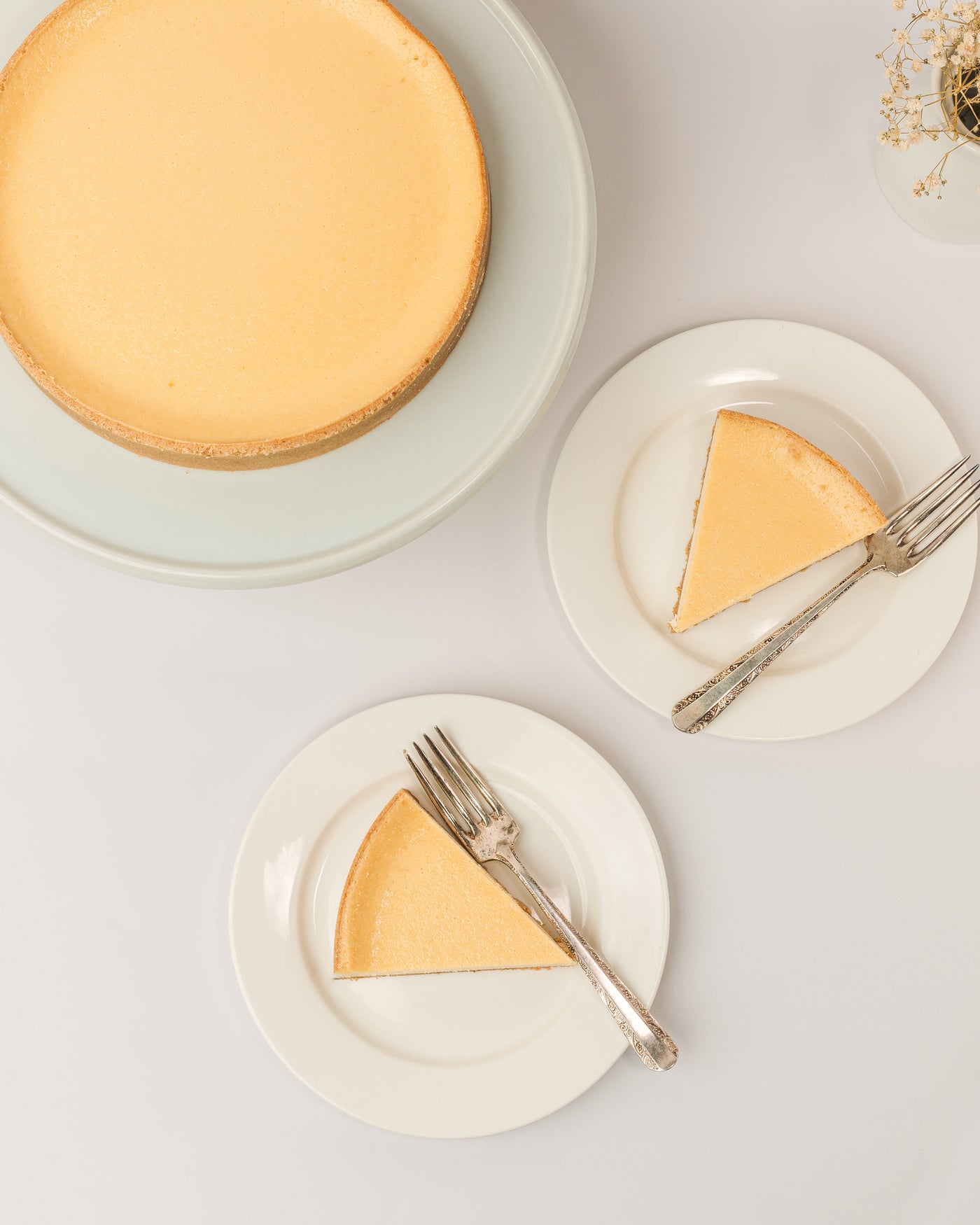 A whole Cheesecake from Loafology Bakery & Café on a cake stand next to two plates, each with a slice of creamy cheesecake and a fork.