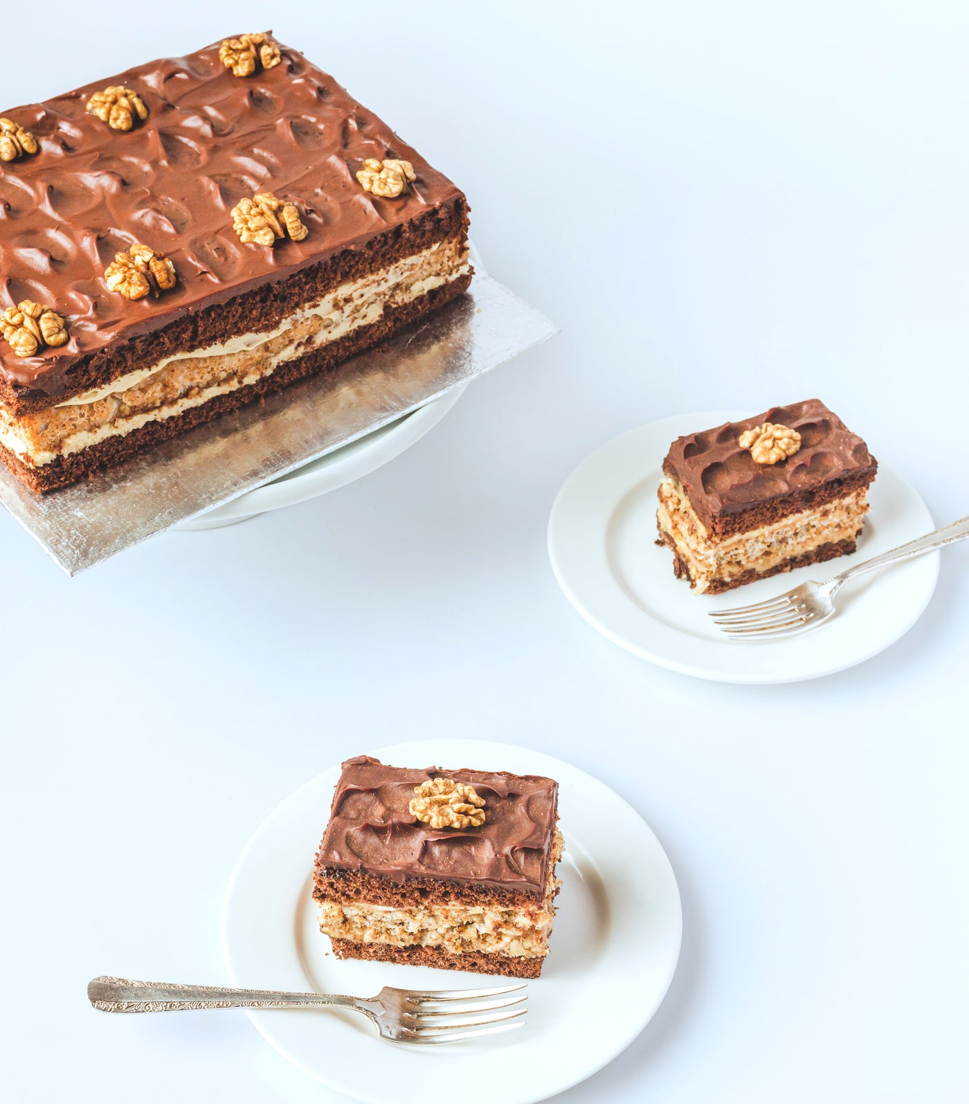 A large rectangular Nikolinka Cake, a chocolate sponge made with organic walnuts, from Loafology Bakery & Café, is presented with two slices on separate white plates, each accompanied by a fork and displayed on a white surface.