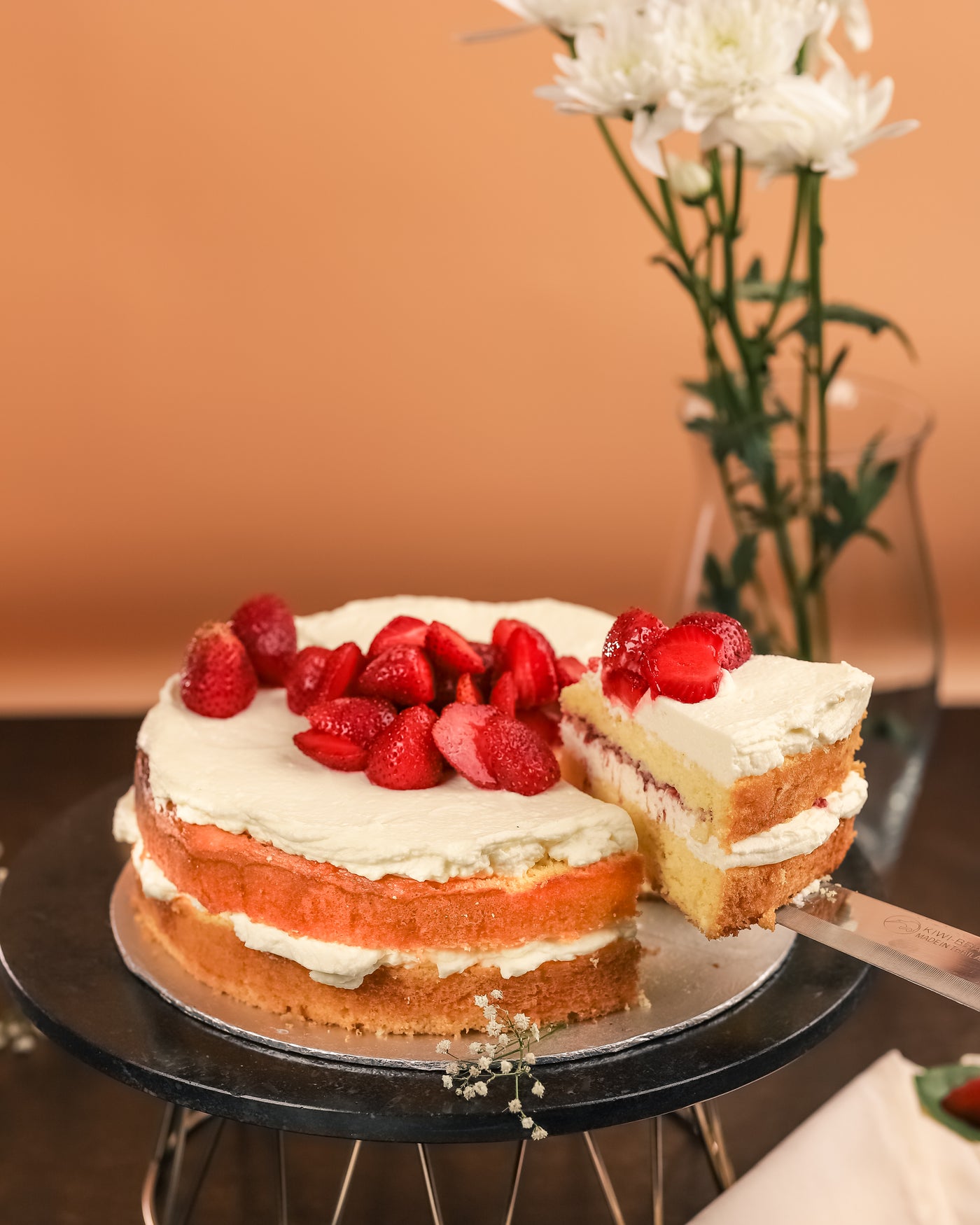 A layered soft Victoria Sponge Cake with whipped French cream sits on a cake stand. A slice is being lifted from the stand with a cake server. A vase with white flowers is in the background, showcasing the delicious offering from Loafology Bakery & Café.