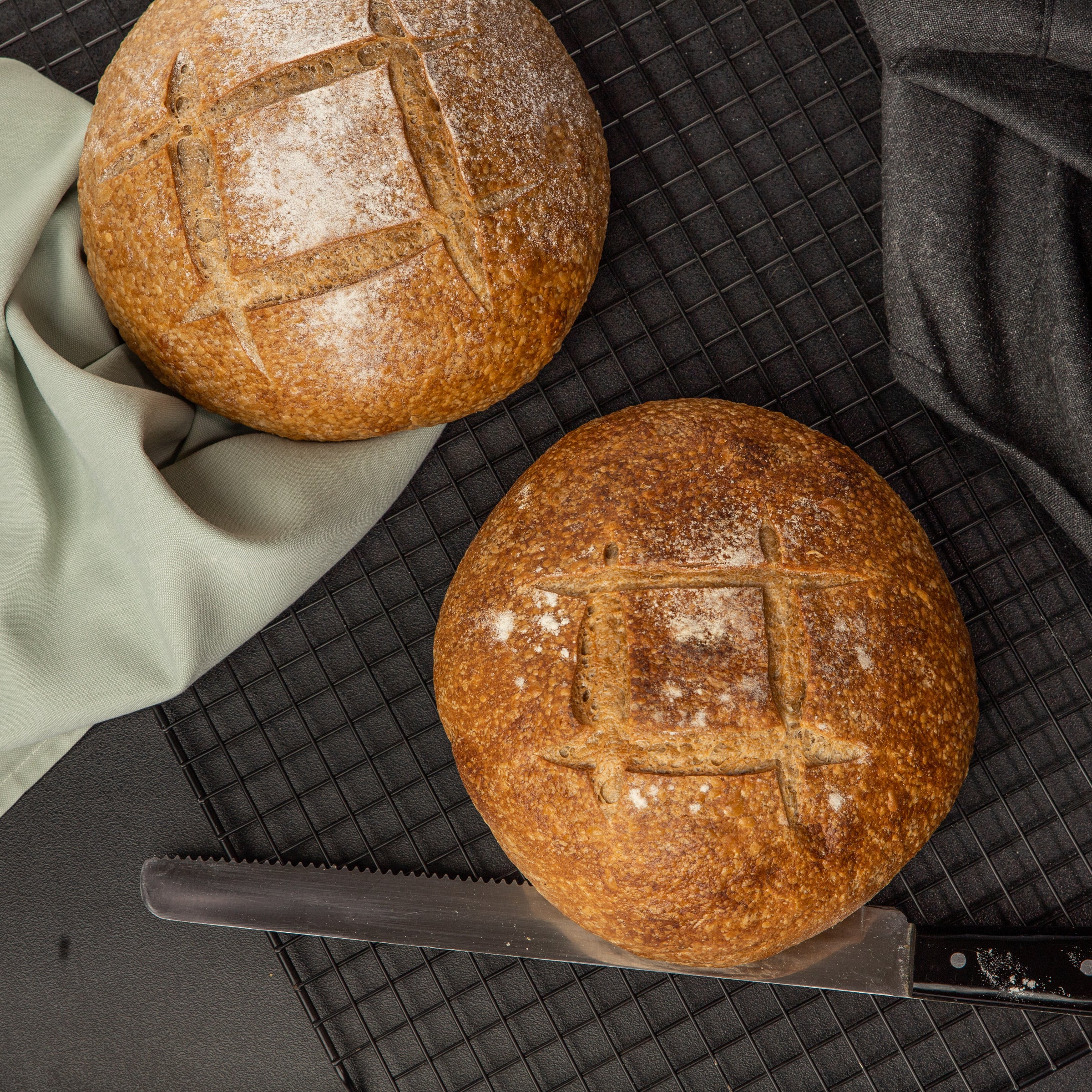 Two round loaves of House bread with scored tops rest on a black wire cooling rack, accompanied by a light green cloth and a black cloth. A serrated knife lies nearby, ready to create delicious sandwiches or make your Breakfast in Bread dreams come true. Enjoy the exquisite quality from Loafology Bakery & Café.