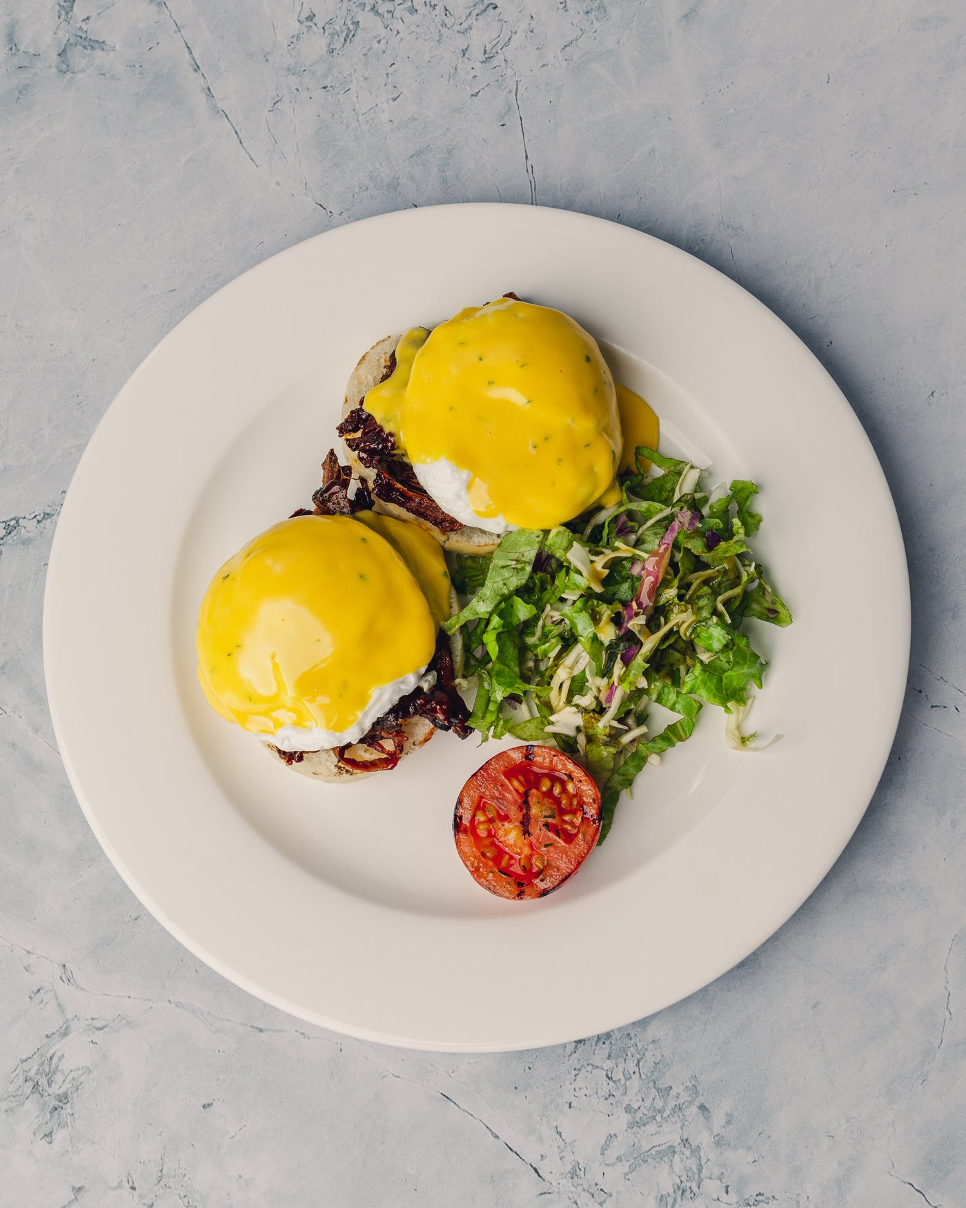 A white plate with Loafology Bakery & Café's Pulled Beef Benedict, featuring two organic poached eggs covered in yellow hollandaise sauce, served with a side of green salad and a grilled tomato half.