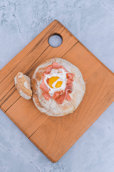 A bread bowl filled with a baked organic egg and slices of Loafology Bakery & Café's smoked salmon sits on a wooden cutting board with a small piece of bread next to it. (Smoked Salmon available on Saturdays and Sundays)