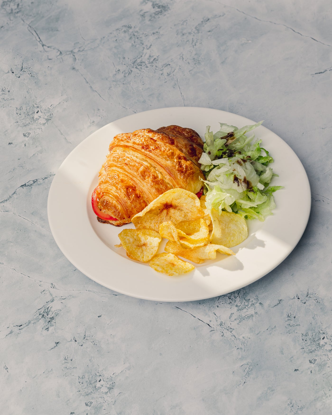 A toasted stuffed croissant from Loafology Bakery & Café, filled with roast chicken and lettuce, served alongside potato chips and a small salad, rests on a white plate placed on a light gray textured surface.