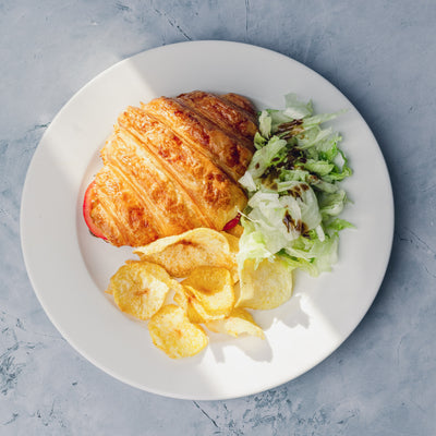 A Toasted Stuffed Croissant filled with roast chicken from Loafology Bakery & Café, accompanied by a side of potato chips and a small serving of lettuce salad, arranged on a gray marble surface.