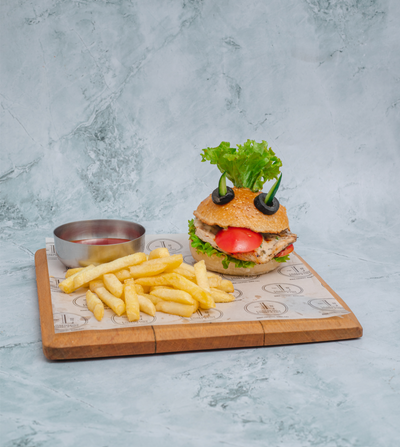 A Loafology Bakery & Café Kids’ Grilled Chicken Burger, complete with lettuce and tomato arranged to resemble a face, is served with French fries and a small container of ketchup on a wooden board.