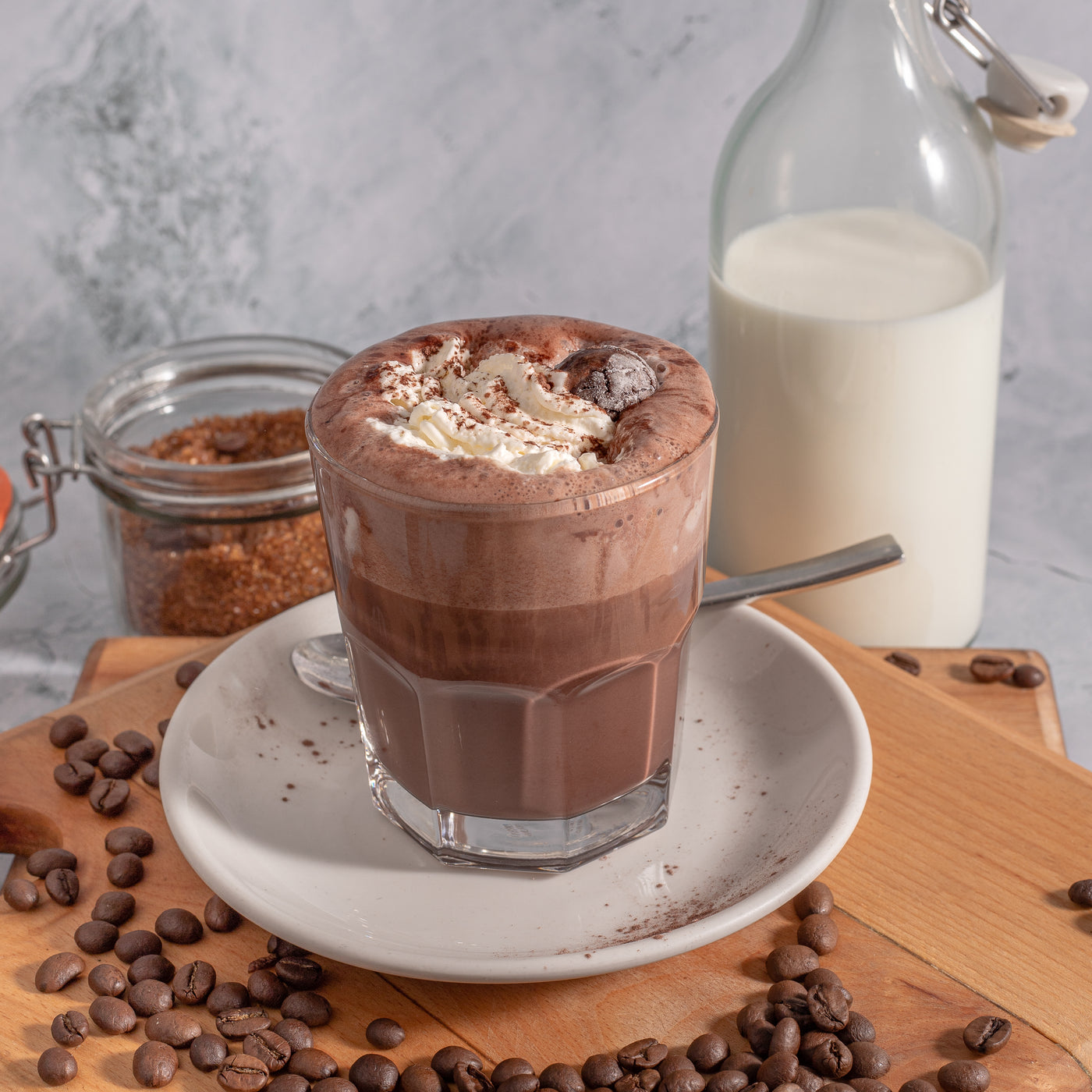 A glass of Vienna Chocolate from Loafology Bakery & Café, topped with whipped cream, served on a white plate alongside a spoon, coffee beans, a jar of brown sugar, and a bottle of milk.