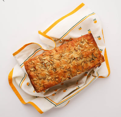 A rectangular loaf of Whole Wheat Sunflower bread from Loafology Bakery & Café, with a crust sprinkled with seeds, displayed on a patterned cloth featuring small orange fruit motifs and a yellow border.