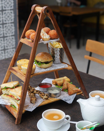 The Loafology Bakery & Café Afternoon Tea Platter features a wooden three-tier stand filled with a variety of snacks, such as mini club sandwiches, a burger, skewers, samosas, fries, pastries, and small desserts. It is accompanied by two teacups with saucers and a teapot brimming with organic loose leaf teas.