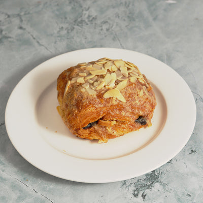 A Loafology Bakery & Café Chocolate Almond Croissant, topped with sliced almonds and featuring a visible chocolate filling, served on a plain white plate placed on a marble surface.