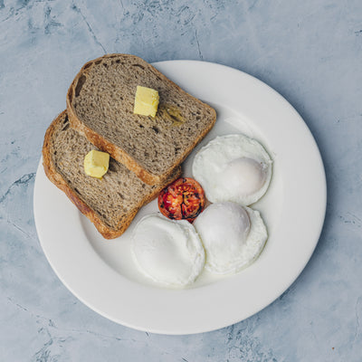 A white plate from Loafology Bakery & Café's "Eggs your style" holds two slices of whole grain bread with butter, two poached eggs, and one slice of roasted tomato, all placed on a light blue stone surface.