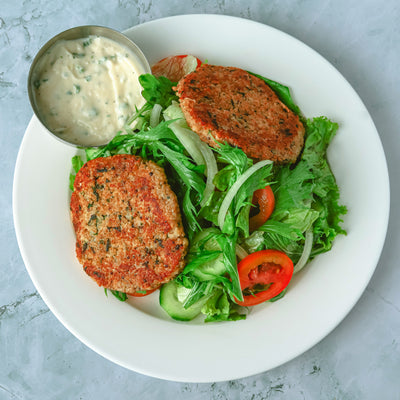 A plate with two quinoa patties on a bed of mixed greens, tomato, cucumber, and onion slices from Loafology Bakery & Café. A small metal cup of white dipping sauce is on the side.