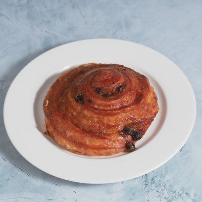 A Raisin Croissant from Loafology Bakery & Café rests on a white plate against a light blue background, displaying its spiral shape, caramelized exterior, and scattered raisins.
